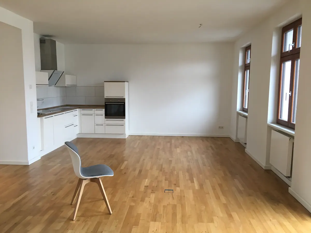 An empty flat with a wood floor, white walls, a fitted kitchen, two windows and one chair